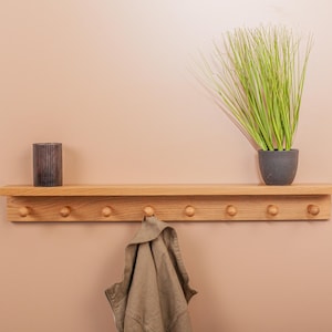 Può includere: Una mensola in legno con pioli, un vaso decorativo e una pianta in vaso con lunghe foglie verdi. Una giacca beige è appesa a un piolo. La mensola è montata su una parete color pesca.
