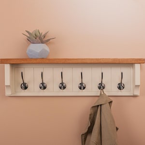 Könnte beinhalten: Eine Wandgarderobe mit sechs Metallhaken und einem Holzregal. Das Regal ist aus hellem Holz gefertigt und die Garderobe ist weiß lackiert. Eine beige Jacke hängt an einem der Haken. Auf dem Regal steht eine dekorative Sukkulente.