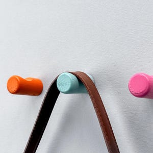 以下が含まれることがあります： オレンジ、水色、ピンクの3色の木製ウォールフック。茶色の革ストラップが水色のフックから吊り下げられています。フックは円筒形で白い壁に取り付けられており、モダンな収納ソリューションを提供します。