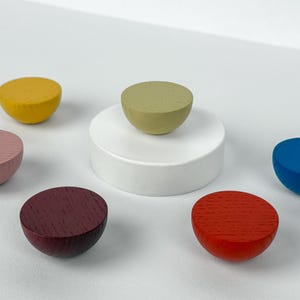 Peut inclure: Ensemble d'objets en bois colorés en forme de demi-sphères. Les couleurs comprennent le jaune, le rose, le bordeaux, le rouge, le bleu et le vert. Une sphère verte repose sur un socle blanc. Les objets sont disposés sur une surface blanche.