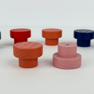 May include: A collection of colorful wooden knobs in various shades, including black, red, orange, navy blue, and light blue. The knobs have a cylindrical shape with a flat top and a slightly wider base. One pink knob is stacked.