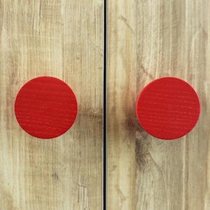 Może przedstawiać: Zbliżenie dwóch czerwonych, okrągłych gałek do szafek na jasnobrązowej drewnianej powierzchni. Gałki są równomiernie rozmieszczone i mają teksturowany wygląd. Widoczne usłojenie drewna.