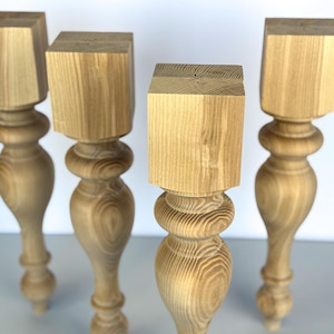 Puede incluir: Cuatro patas de mesa de madera sin terminar. Cada pata tiene una parte superior cuadrada, una sección media bulbosa y torneada, y una base cónica. La veta de la madera es visible y las patas son de color marrón claro.
