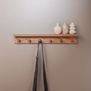 May include: A wooden shelf with six copper-colored hooks, holding three decorative vases. A black crossbody bag hangs from one hook. The shelf is mounted on a neutral-toned wall, creating a minimalist aesthetic.