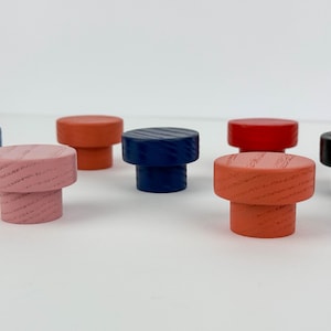 Pode incluir: Uma coleção de puxadores de madeira coloridos em várias tonalidades, incluindo azul, rosa, laranja, vermelho e preto. Cada puxador tem uma base cilíndrica e um topo plano e redondo, mostrando o grão da madeira. Dispostos sobre uma superfície branca.