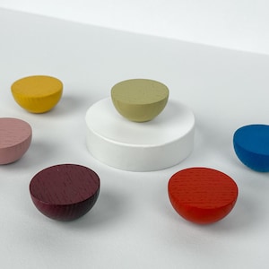 Könnte beinhalten: Eine Sammlung farbenfroher, halbkugelförmiger Holzobjekte. Die Farben umfassen Gelb, Rosa, Burgunderrot, Rot, Blau und Grün. Eine grüne Kugel ruht auf einem weißen Sockel. Die Objekte sind auf einer weißen Oberfläche angeordnet.