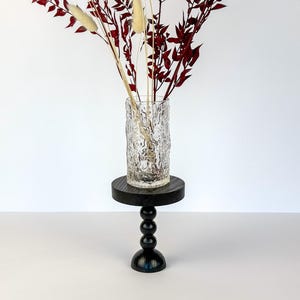 May include: A clear glass vase with a textured surface, holding dried red and beige decorative stems, sits atop a small, black, wooden pedestal. The pedestal has a round top and a base with a series of stacked spheres.