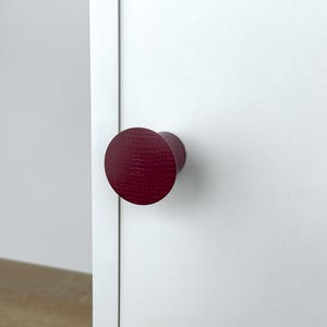 Może przedstawiać: Bordowy uchwyt do szafki z teksturowaną, okrągłą główką przymocowany do białych drzwi. Uchwyt ma cylindryczną podstawę. Drzwi są białe, co stanowi kontrast. Tekstura uchwytu dodaje wizualnego zainteresowania.