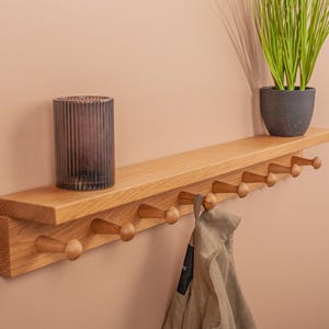 May include: A wooden wall shelf with a row of coat hooks. A gray glass candle holder and a potted plant sit on the shelf. A tan jacket hangs from one of the hooks. The shelf is mounted on a pink wall.