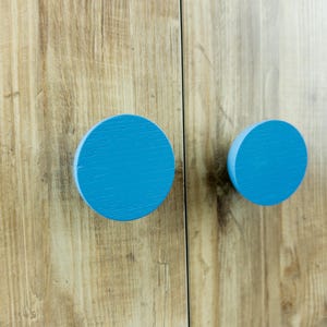 May include: Close-up of two round, light blue cabinet knobs against a light brown wood-grain surface. The knobs are evenly spaced and appear to be made of wood. The wood grain has a natural, textured appearance.