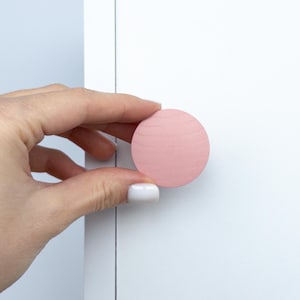 May include: A round, pale pink wooden cabinet knob. The knob is held against a white cabinet door. The knob has a smooth, matte finish and a visible wood grain pattern. The knob is a simple, modern design.