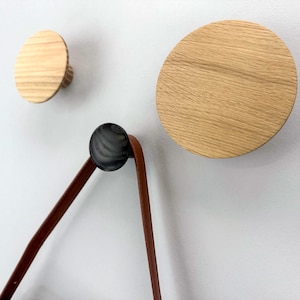 Könnte beinhalten: Drei Wandhaken aus Holz in verschiedenen Größen. Zwei sind kreisförmig, einer groß und einer klein, aus hellem Holz. Ein dritter Haken ist schwarz und hält einen braunen Lederriemen.