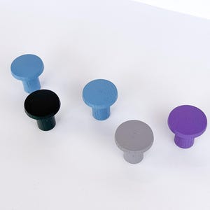Pode incluir: Um conjunto de seis puxadores de armário de madeira em várias cores: azul claro, preto, azul médio, cinza e roxo. Cada puxador tem um topo redondo e uma base cilíndrica, perfeitos para adicionar cor aos móveis.