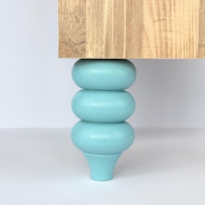 May include: A wooden table with a light brown finish, supported by a single, turquoise-painted, decorative leg. The leg has a stacked, rounded design. The table leg adds a pop of colour and a unique aesthetic to the furniture.