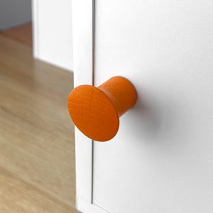 以下が含まれることがあります： オレンジ色の木製キャビネットノブ。円筒形の本体と円形の頭部があります。ノブは白いキャビネットドアに取り付けられています。木目はノブの表面に見えます。背景には木の床とドアの様子が写っています。
