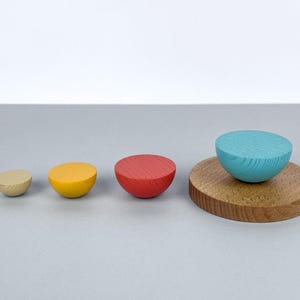 Puede incluir: Un conjunto de objetos de madera en forma de media esfera de varios colores. Hay una media esfera beige, amarilla y roja, con una media esfera turquesa más grande sobre un disco de madera. Los objetos están dispuestos sobre una superficie gris claro.