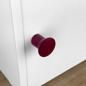 May include: A close-up of a white cabinet with a dark burgundy, cylindrical knob. The knob has a flat, circular face. The cabinet is made of white wood and has a simple, modern design. The knob is the focal point.