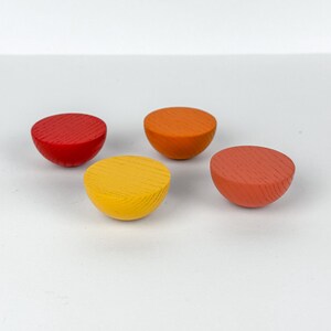 May include: Four colorful, half-sphere wooden objects in red, orange, yellow, and coral. Each has a flat, textured surface. The objects are arranged on a white surface, creating a simple, yet vibrant composition.