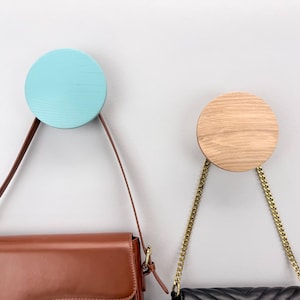 May include: Two wooden wall hooks, one aqua and one natural wood, each with a leather or chain strap holding a purse. The aqua hook holds a brown purse, and the wood hook holds a black purse.