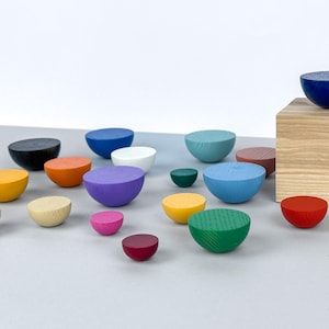 May include: A collection of colourful wooden half-sphere ornaments in various sizes and colours, including pink, red, green, blue, yellow, orange, and black. The ornaments are arranged on a light grey surface.