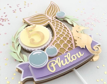 May include: A mermaid-themed cake topper with a gold glitter tail, seahorse, and the number 5. The topper includes a banner with the name "Philou" in gold lettering, surrounded by bubbles and greenery. The color palette includes gold, pink, lavender, and light blue.