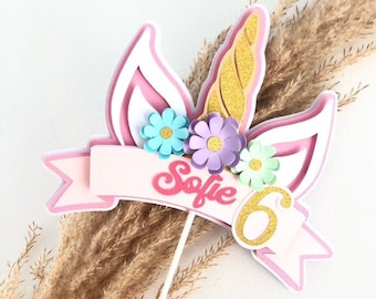 May include: Pink unicorn cake topper with a gold horn, white ears, and colorful flowers. The topper has a banner with the name "Sofie" and the number "6" on it.