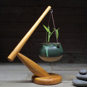 May include: A wooden plant stand with a long arm that holds a green ceramic pot with a small plant hanging from it. The stand has a round base and is made of natural wood. There are three grey stones stacked on the table in the background.