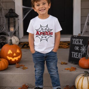Peut inclure: Un t-shirt blanc &agrave; manches courtes orn&eacute; d'un motif de toile d'araign&eacute;e noire avec une petite araign&eacute;e et le pr&eacute;nom Archie imprim&eacute; en lettres rouges au centre. Le t-shirt est port&eacute; avec un jean bleu et des baskets noires &agrave; semelles blanches.
