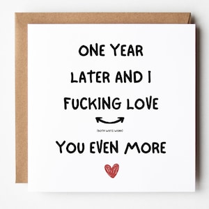 Op de afbeelding: Vierkante wenskaart met een bruine envelop. De kaart heeft zwarte tekst die luidt: "EEN JAAR LATER EN IK FUCKING LOVE YOU EVEN MORE" met een rood hart en een smiley-emoji.