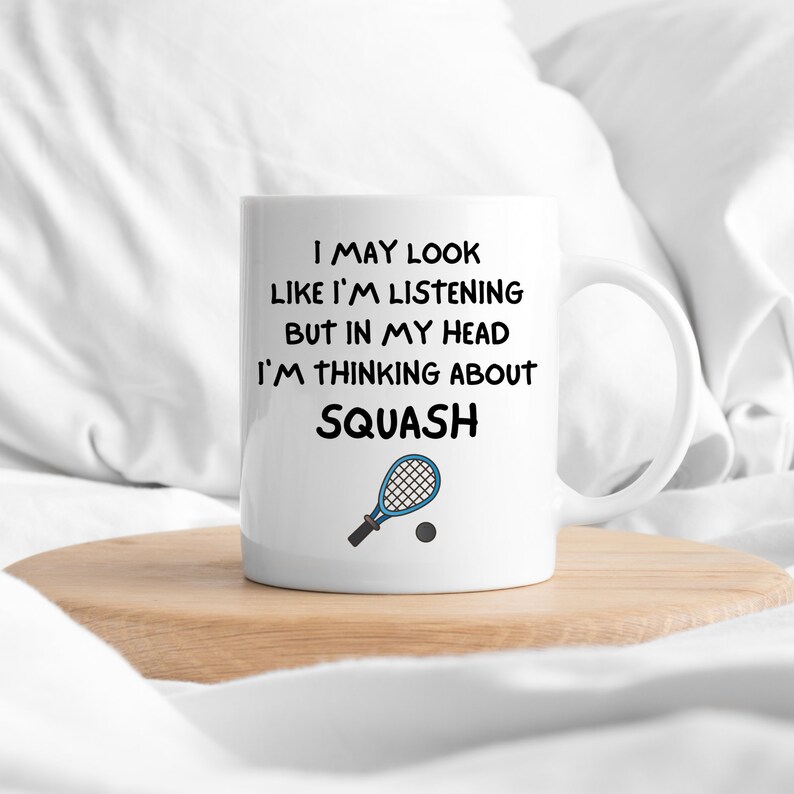 May include: White ceramic mug with a black and blue design. The mug features the text "I may look like I'm listening but in my head I'm thinking about SQUASH" and a cartoon image of a squash racket and ball.