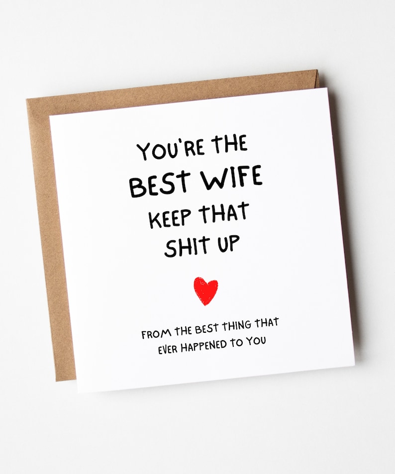 以下が含まれることがあります： 黒い文字で「YOU'RE THE BEST WIFE KEEP THAT SHIT UP」と書かれた正方形の白いグリーティングカード。テキストの下には小さな赤いハートがあり、その下に「FROM THE BEST THING THAT EVER HAPPENED TO YOU」と書かれています。カードは茶色の封筒の上に置かれています。