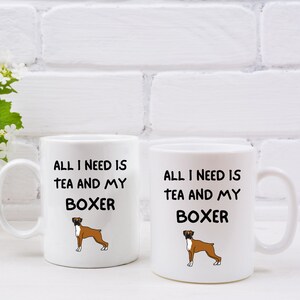 May include: Two white ceramic mugs with black text and a cartoon boxer dog. The text on the mugs reads "All I need is tea and my boxer".