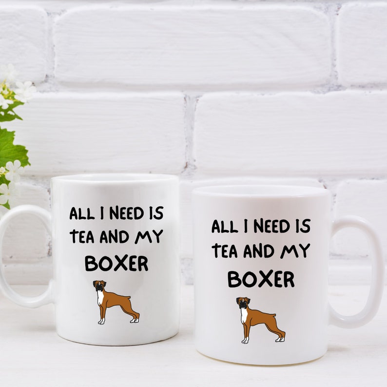 May include: Two white ceramic mugs with black text and a cartoon boxer dog. The text on the mugs reads "All I need is tea and my boxer".