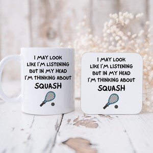 May include: White mug and coaster with a blue squash racket and ball graphic. The text reads "I may look like I'm listening but in my head I'm thinking about squash."