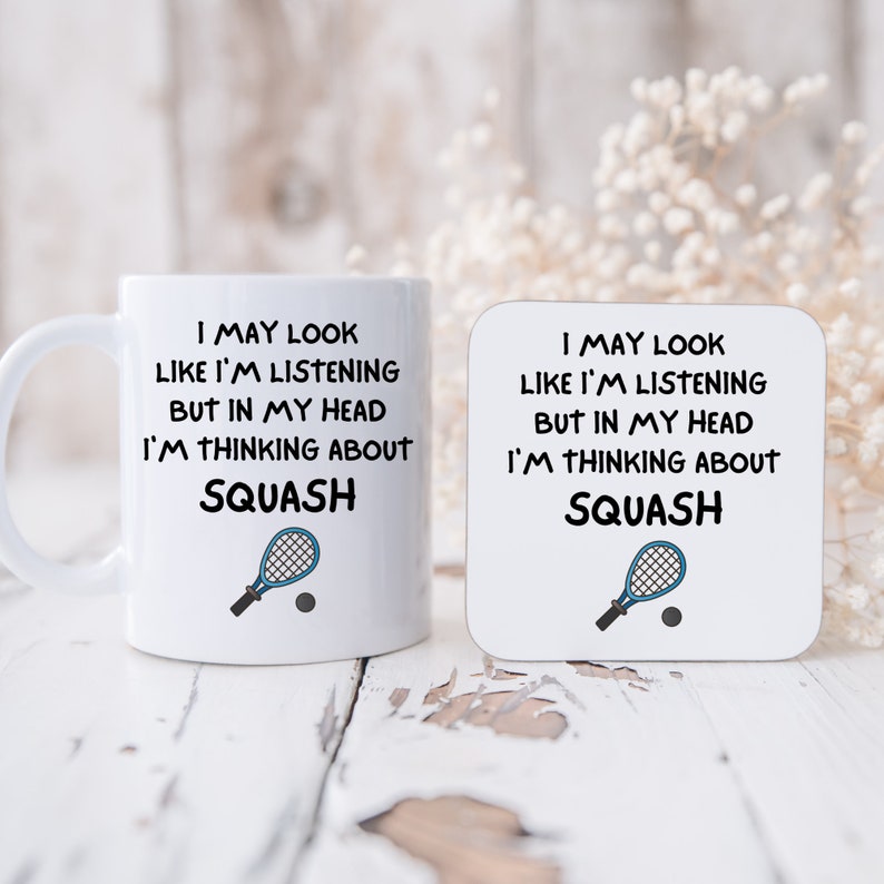 May include: White mug and coaster with a blue squash racket and ball graphic. The text reads "I may look like I'm listening but in my head I'm thinking about squash."
