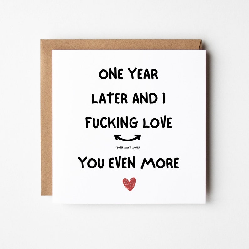 Op de afbeelding: Vierkante wenskaart met een bruine envelop. De kaart heeft zwarte tekst die luidt: "EEN JAAR LATER EN IK FUCKING LOVE YOU EVEN MORE" met een rood hart en een smiley-emoji.