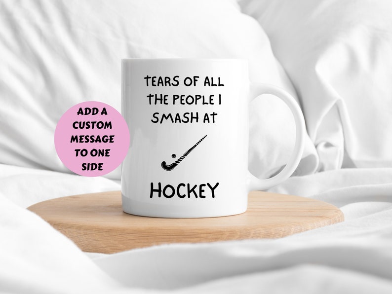 May include: White ceramic coffee mug with black text that reads "Tears of all the people I smash at Hockey" and a black hockey stick graphic.  The mug is on a wooden coaster.