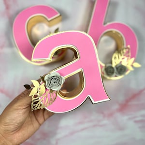 Può includere: Lettere decorative rosa e oro, inclusa la lettera "A", con accenti floreali. Le lettere hanno un interno dorato e sono decorate con fiori di carta e foglie d'oro. La lettera "A" è tenuta in mano.
