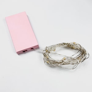 Puede incluir: Un paquete de baterías rosa con un cable conectado a una cadena de luces LED blancas cálidas. Las luces están enrolladas.