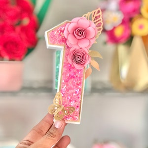 Puede incluir: Un número uno decorativo, con purpurina rosa y flores de papel. El número está adornado con una mariposa dorada y detalles de hojas doradas. El número mide aproximadamente 15 cm de alto, ideal para una fiesta de cumpleaños.