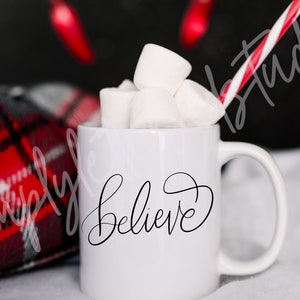 Könnte beinhalten: Eine weiße Keramiktasse mit dem Wort "Believe" in schwarzer Kursivschrift. Die Tasse ist mit Marshmallows gefüllt und hat einen rot-weiß gestreiften Strohhalm.