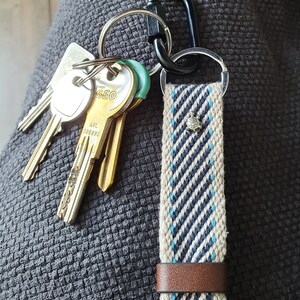 Peut inclure: Un porte-cl&eacute;s avec une sangle tiss&eacute;e beige et bleu marine &agrave; rayures diagonales, orn&eacute;e d'un petit charm argent&eacute; et d'une bande en cuir marron. Plusieurs cl&eacute;s et un mousqueton noir sont attach&eacute;s.