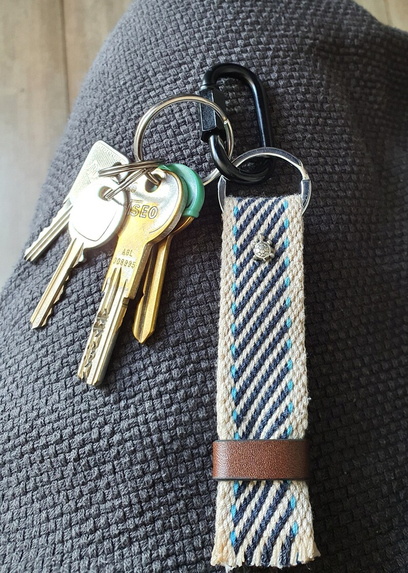 Peut inclure: Un porte-cl&eacute;s avec une sangle tiss&eacute;e beige et bleu marine &agrave; rayures diagonales, orn&eacute;e d'un petit charm argent&eacute; et d'une bande en cuir marron. Plusieurs cl&eacute;s et un mousqueton noir sont attach&eacute;s.