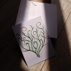 Peut inclure: Une carte de vœux blanche avec un dessin à la main de branches stylisées en noir, vert et bleu. La carte est sur une enveloppe blanche, avec un article en cuir marron en arrière-plan. L'image est éclairée par un rayon de soleil.