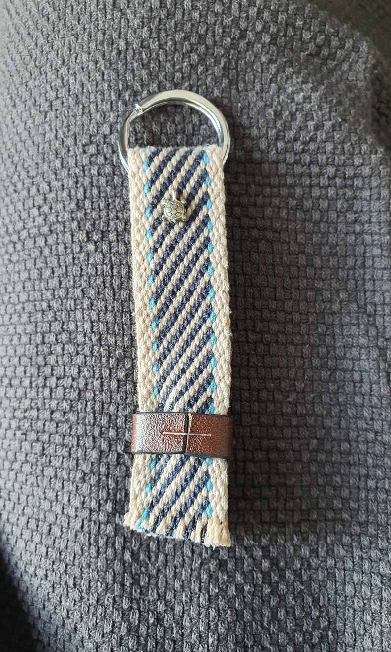 Peut inclure: Porte-cl&eacute;s tiss&eacute; avec un anneau argent&eacute;. La sangle pr&eacute;sente un fond cr&egrave;me avec des rayures diagonales bleu marine et bleu clair. Un petit charme de tortue argent&eacute; est attach&eacute;, et une bande de cuir marron s&eacute;curise la sangle.