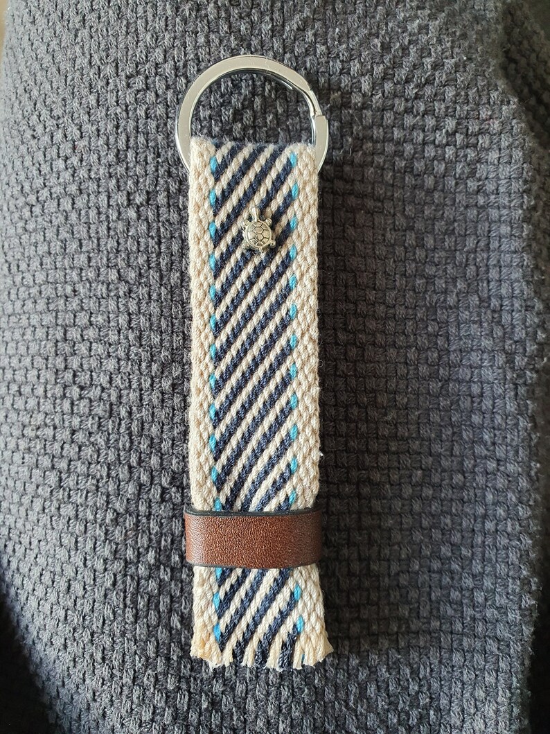 Peut inclure: Un porte-cl&eacute;s tiss&eacute; avec un motif ray&eacute; cr&egrave;me, marine et bleu clair. Il est orn&eacute; d'un petit charm en forme de tortue argent&eacute;e, d'une bande en cuir marron et d'un porte-cl&eacute;s argent&eacute;.