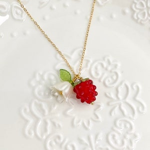 May include: A gold chain necklace with a pendant depicting a red raspberry, white flower, and green leaf. The necklace is presented on a white, decorative plate. Two red raspberry beads are also visible.
