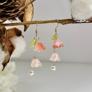 May include: A pair of dangle earrings with pink and white flower-shaped charms and a pearl accent. The earrings are hanging from a branch with cotton balls.