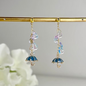 Peut inclure: Une paire de boucles d'oreilles en or avec des parapluies bleus et des perles de cristal irisées qui pendent.