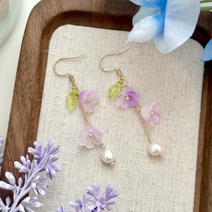 May include: A pair of dangle earrings featuring a delicate gold chain and three purple flower charms. Each earring is finished with a single white pearl.